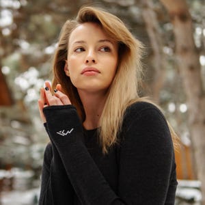 May include: A dark gray long-sleeved shirt with a textured pattern. The shirt features a white embroidered mountain logo on the left wrist. The person in the image is wearing the shirt outdoors.