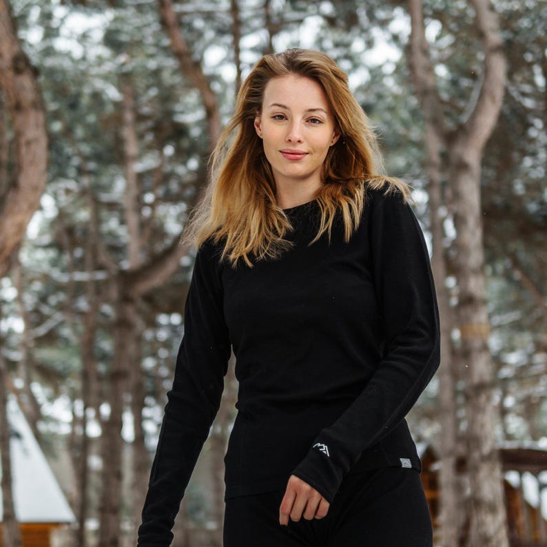 May include: A person wearing a black long-sleeved shirt and black pants. The person is standing in a snowy forest. The shirt has a small white logo on the sleeve.