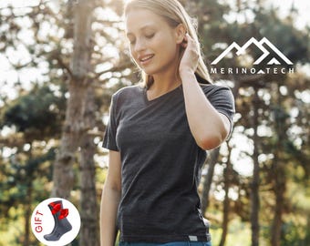 May include: A dark charcoal gray V-neck t-shirt with short sleeves and a small logo tag at the hem. A circular inset shows a gray sock with a red bow and the text GIFT. The logo MERINO TECH with a mountain graphic is visible in the upper right.