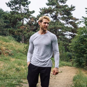 May include: A light gray long-sleeve shirt and black pants. The shirt has a crew neck and a slim fit. The person is walking on a path in a wooded area. The shirt is made of a soft, comfortable material.