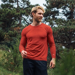 May include: A long-sleeved, rust-colored shirt. The shirt is made of a soft, textured material. The person in the image is wearing black pants. The background features green trees and foliage.