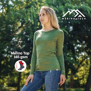 May include: A long-sleeve olive green crew neck top with a heathered texture. The garment features the text Merino Top 165 gsm and a circular graphic showing a gray sock with red accents. The brand logo Merino Tech is visible in the upper corner.