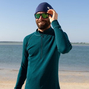 May include: A teal long-sleeve shirt with a quarter-zip neckline, paired with a navy blue beanie and black sunglasses. The sunglasses have green reflective lenses. The person is smiling outdoors near the water.