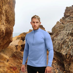 May include: A light blue long-sleeve shirt with a quarter-zip collar. The shirt is made of a soft, textured material. The model is standing in front of a rocky background near the ocean. The shirt is designed for outdoor activities.