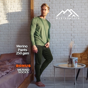 May include: A long-sleeve olive green shirt and dark olive green pants. The text on the image reads Merino Tech, Merino Pants 250 gsm, and BONUS MERINO SOCKS. The clothing is worn by a person standing in a room with a white brick wall and a side table.