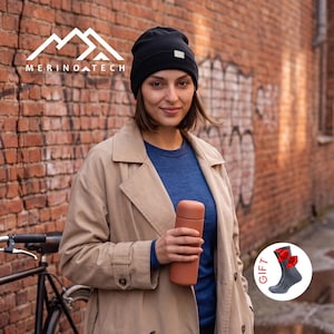 May include: A black beanie with a small white tag, a blue long-sleeve shirt, and a tan trench coat. A matte terracotta-colored insulated bottle is held in the hand. A circular graphic shows a gray sock with a red bow and the text GIFT.