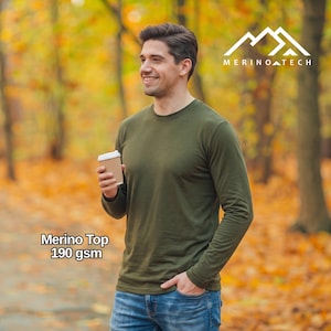 Può includere: Una maglia a maniche lunghe con girocollo di colore verde oliva con la scritta Merino Top 190 gsm stampata in basso a sinistra. La maglia presenta il logo Merino Tech nell'angolo in alto a destra. Il capo è abbinato a jeans blu e un bicchiere di carta.