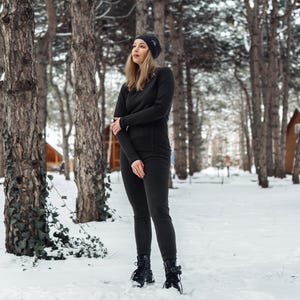 K&ouml;nnte beinhalten: Eine Person in einem schwarzen Thermotop, Leggings und einer M&uuml;tze in einem verschneiten Wald. Das Outfit umfasst ein lang&auml;rmliges Oberteil, eng anliegende Leggings und eine M&uuml;tze. Die Person tr&auml;gt schwarze Stiefel mit Schn&uuml;rsenkeln.