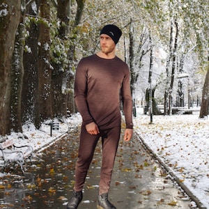 May include: A brown thermal base layer set, including a long-sleeved shirt and pants, is worn with a black beanie. The outfit is designed for cold weather. The person is standing on a path in a snowy park.