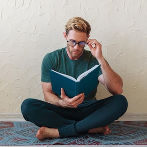 May include: A person with blonde hair wearing a dark green t-shirt and dark pants, sitting cross-legged on a patterned mat while reading a dark blue hardcover book. The person is wearing black-rimmed glasses and holding the book open with both hands.