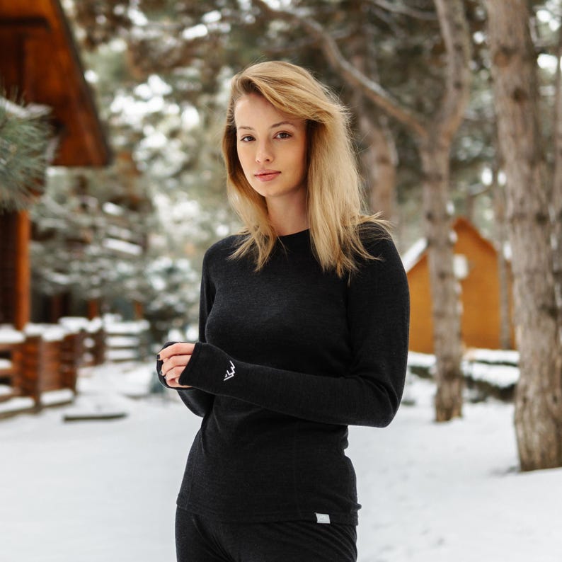 May include: A dark gray long-sleeved shirt and matching pants. The shirt has a crew neck and a small white logo on the cuff. The outfit is made of a soft, warm material, perfect for cold weather. The background is a snowy outdoor scene.