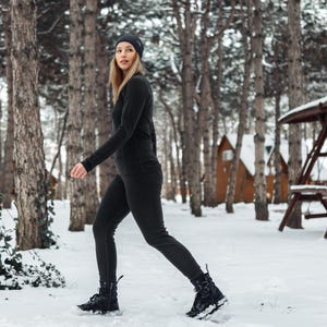 K&ouml;nnte beinhalten: Eine Person in einem schwarzen Thermotop, Leggings und einer M&uuml;tze geht durch einen verschneiten Wald. Das Outfit umfasst schwarze Stiefel. Der Hintergrund zeigt schneebedeckten Boden, B&auml;ume und Holzstrukturen.
