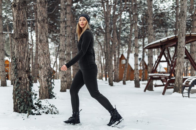 K&ouml;nnte beinhalten: Eine Person in einem schwarzen Thermotop, Leggings und einer M&uuml;tze geht durch einen verschneiten Wald. Das Outfit umfasst schwarze Stiefel. Der Hintergrund zeigt schneebedeckten Boden, B&auml;ume und Holzstrukturen.