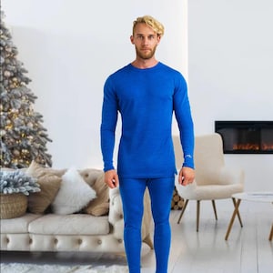 May include: A person wearing a blue thermal base layer set. The set includes a long-sleeved crew neck shirt and matching pants. The clothing is a solid, vibrant blue color, and appears to be made of a soft, textured material. The person is standing in a well-lit room.
