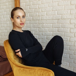 May include: A long-sleeved black crewneck shirt and matching black pants. The shirt features a small white embroidered logo on the left cuff. The fabric appears to be a solid, opaque black material with a smooth texture and a fitted silhouette.
