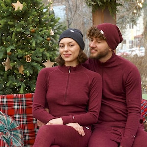 May include: A couple wearing matching burgundy thermal tops and leggings, with a dark gray beanie and burgundy beanie. The tops have a quarter-zip closure. They are sitting in front of a decorated Christmas tree.