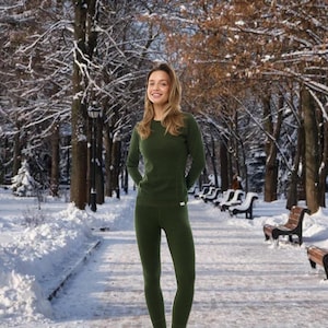 May include: A person wearing a dark green long-sleeved shirt and matching leggings. The outfit is made of a soft, possibly thermal material. The person is standing in a snowy park with trees and benches in the background.