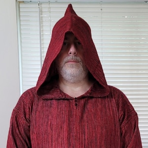 May include: A red hooded tunic with a black and red striped pattern. The tunic has two pockets and a zipper closure. The person is wearing gray pants.