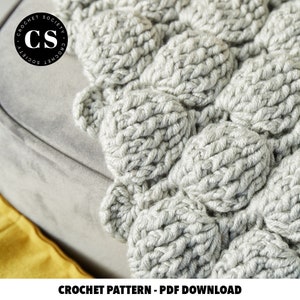 Pode incluir: Um close-up de um cobertor de croch&ecirc; cinza com um padr&atilde;o texturizado em forma de bolhas. O cobertor &eacute; feito com um fio grosso e tem um toque macio e aconchegante.
