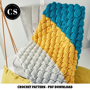 Pode incluir: Uma manta de croch&ecirc; com um padr&atilde;o de ponto texturizado e inchado em tr&ecirc;s cores: cinza, amarelo e azul-petr&oacute;leo. A manta est&aacute; drapeada sobre uma cadeira com uma almofada amarela em primeiro plano. O texto "CROCHET PATTERN - PDF DOWNLOAD" est&aacute; na parte inferior da imagem.