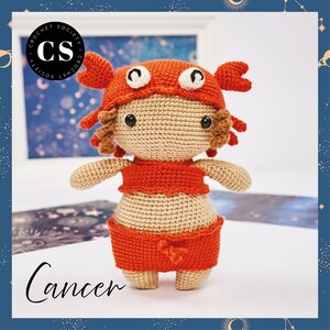 Puede incluir: Una muñeca de crochet que lleva un disfraz de cangrejo rojo. La muñeca tiene el pelo castaño y lleva un atuendo rojo y beige. La muñeca está de pie sobre una superficie blanca. El texto "Cáncer" está escrito debajo de la muñeca.
