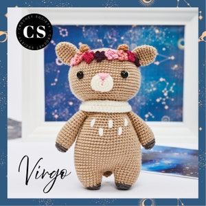May include: A crocheted deer with a pink and purple flower crown, white scarf, and brown and white spots. The deer is standing on a white surface and the background is a blue sky with stars. The text "Virgo" is written in black cursive below the deer.
