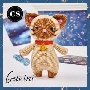 May include: Crocheted cat amigurumi toy with a brown and beige color scheme. The cat is wearing a red collar with a gold bell. The cat is holding a blue fish toy. The cat is labeled "Gemini".