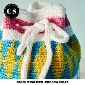 Pu&ograve; includere: Una borsa all'uncinetto con un motivo a righe bianche, rosa e blu. La borsa ha un cordoncino bianco e un motivo geometrico giallo e blu sul fondo.