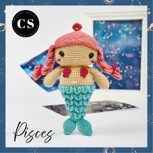 Könnte beinhalten: Eine gehäkelte Meerjungfrauenpuppe mit rosa Haaren, einem beigefarbenen Oberkörper und einem türkisfarbenen Schwanz. Die Puppe steht auf einer weißen Oberfläche und der Text "Pisces" ist in Schwarz unter der Puppe geschrieben.