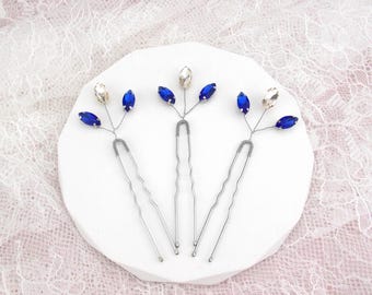May include: Three silver hair pins with blue and clear crystal accents. The pins are arranged on a white surface.