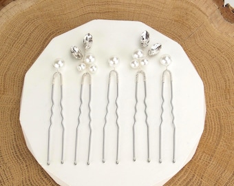 Puede incluir: Conjunto de seis horquillas plateadas. Cada horquilla presenta un adorno de perlas y cristales. Las horquillas están dispuestas sobre una superficie geométrica blanca, sobre un fondo de madera natural. Ideal para bodas u ocasiones especiales.