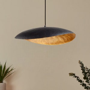 Peut inclure: Un luminaire suspendu moderne, noir et doré. La lampe a un extérieur gris foncé texturé et un intérieur doré. Elle est suspendue à un cordon noir, éclairant une table à manger avec des assiettes, une tasse et un vase avec de la verdure.