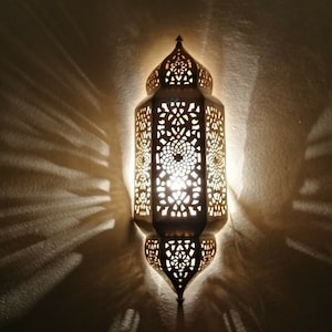 Könnte beinhalten: Verzierte, goldfarbene Wandleuchte mit aufwendigen Ausschnittmustern, die dekorative Licht- und Schattenmuster an die Wand wirft. Die Lampe hat eine spitze Ober- und Unterseite, und die Lichtquelle ist durch das offene Design sichtbar.