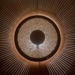Op de afbeelding: Een ronde, witte plafondlamp met een decoratief, ingewikkeld patroon van uitsparingen. Het licht werpt een schaduw van stralende lijnen op het plafond.