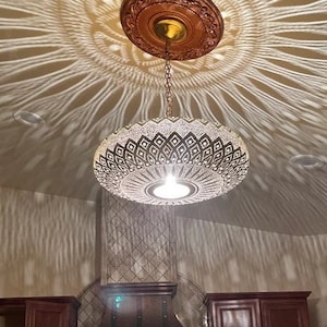 Puede incluir: Una gran l&aacute;mpara de techo de metal dorado adornada con un patr&oacute;n geom&eacute;trico cuelga de una cadena en una cocina. La l&aacute;mpara proyecta una sombra decorativa en el techo. La cocina tiene gabinetes marrones y una campana de cocina de cobre con un salpicadero de mosaico que presenta un dise&ntilde;o de &aacute;rbol de la vida.