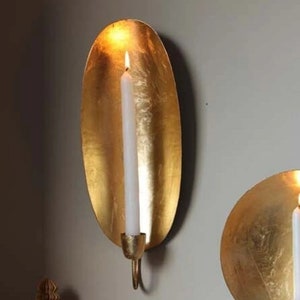 May include: Two gold metal wall candle holders. The holder on the left is oval shaped and has a white candle inside. The holder on the right is round.