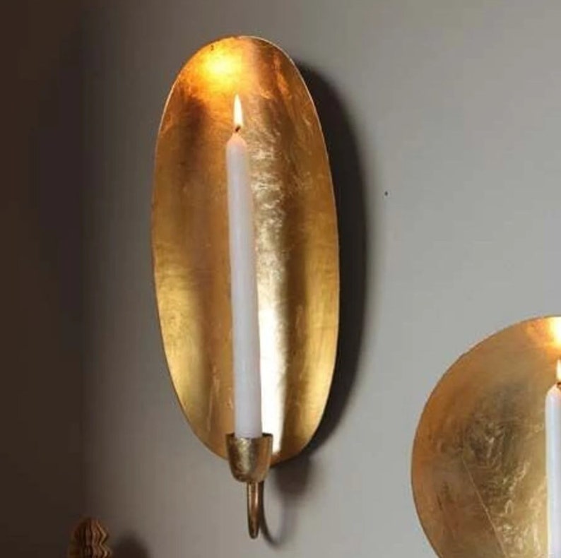 May include: Two gold metal wall candle holders. The holder on the left is oval shaped and has a white candle inside. The holder on the right is round.