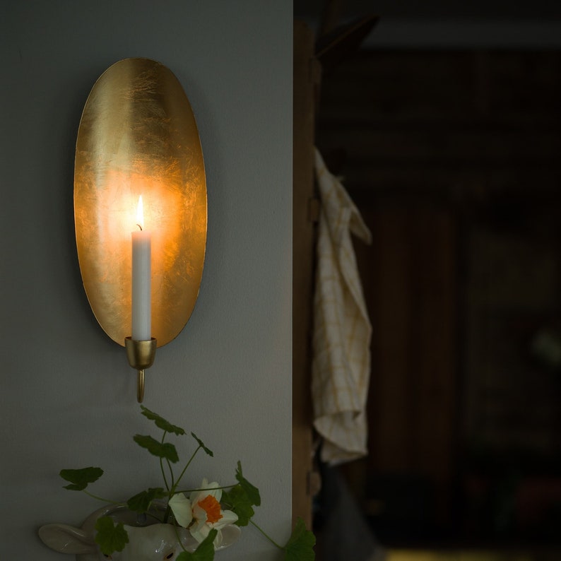 May include: A gold metal wall sconce with an oval-shaped backplate and a candle holder. A white candle is lit inside the sconce, casting a warm glow.