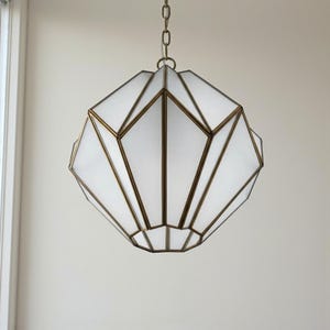 May include: Geometric pendant light with a brass frame and white glass panels. The fixture is suspended from a chain and features a multifaceted design, creating a modern and elegant aesthetic. The light is suitable for interior spaces.