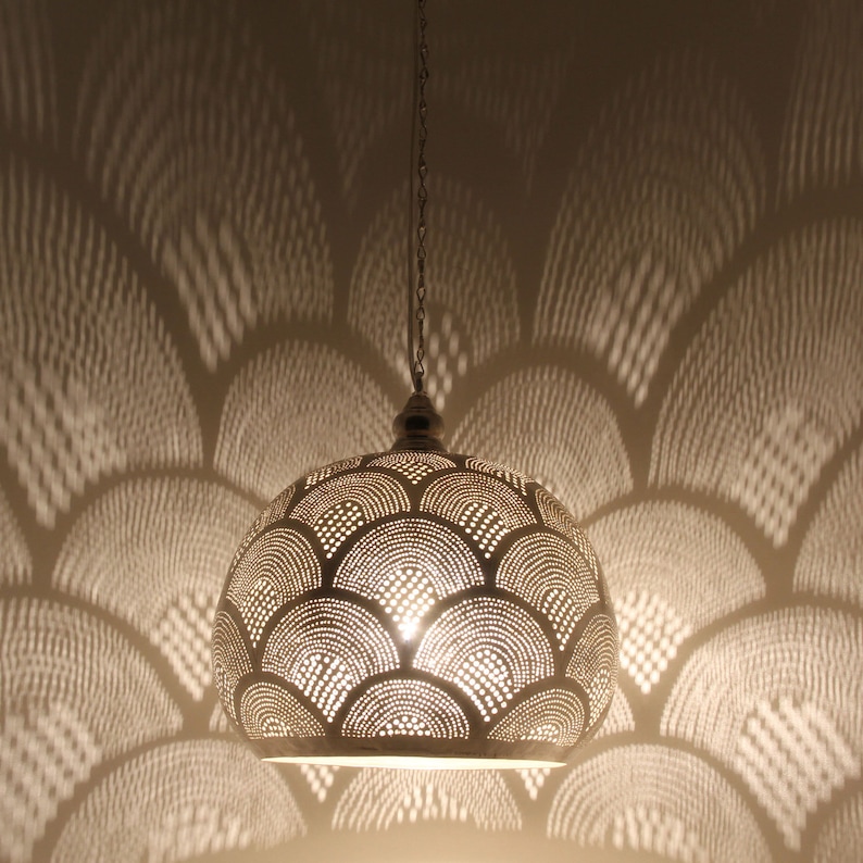 May include: A dome-shaped metal pendant light fixture featuring a perforated pattern of repeating semicircles. The light shines through the small holes, casting a large, intricate, and glowing scalloped shadow pattern onto the surrounding wall surface.