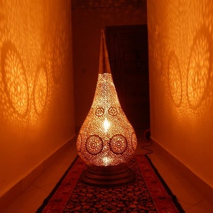 K&ouml;nnte beinhalten: Eine tropfenf&ouml;rmige Bodenlampe aus Metall mit einem komplizierten Lochmuster, das kreisf&ouml;rmige Lichtprojektionen an die W&auml;nde wirft. Die Lampe strahlt ein warmes, goldenes Licht aus und hat eine spitze Oberseite sowie einen runden Sockel.