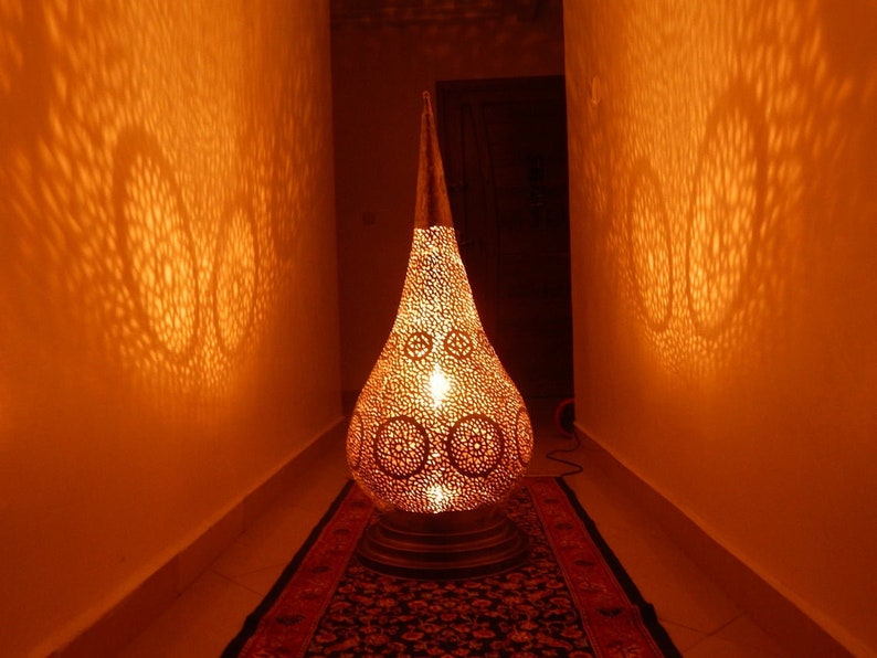 K&ouml;nnte beinhalten: Eine tropfenf&ouml;rmige Bodenlampe aus Metall mit einem komplizierten Lochmuster, das kreisf&ouml;rmige Lichtprojektionen an die W&auml;nde wirft. Die Lampe strahlt ein warmes, goldenes Licht aus und hat eine spitze Oberseite sowie einen runden Sockel.