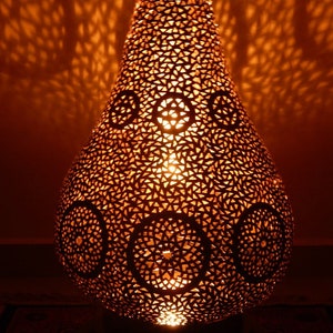 K&ouml;nnte beinhalten: Eine birnenf&ouml;rmige Metalllampe mit einem warmen orangefarbenen Lichtschein. Die Oberfl&auml;che weist komplexe, lasergeschnittene geometrische Muster auf, darunter sich wiederholende Dreiecksformen und kreisf&ouml;rmige Medaillons, die detaillierte Schatten werfen.