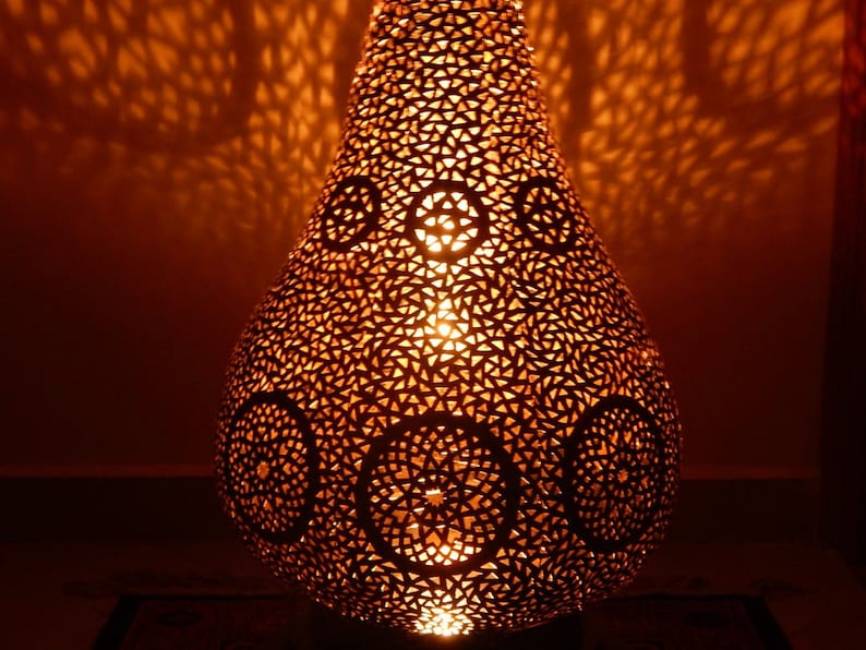 K&ouml;nnte beinhalten: Eine birnenf&ouml;rmige Metalllampe mit einem warmen orangefarbenen Lichtschein. Die Oberfl&auml;che weist komplexe, lasergeschnittene geometrische Muster auf, darunter sich wiederholende Dreiecksformen und kreisf&ouml;rmige Medaillons, die detaillierte Schatten werfen.
