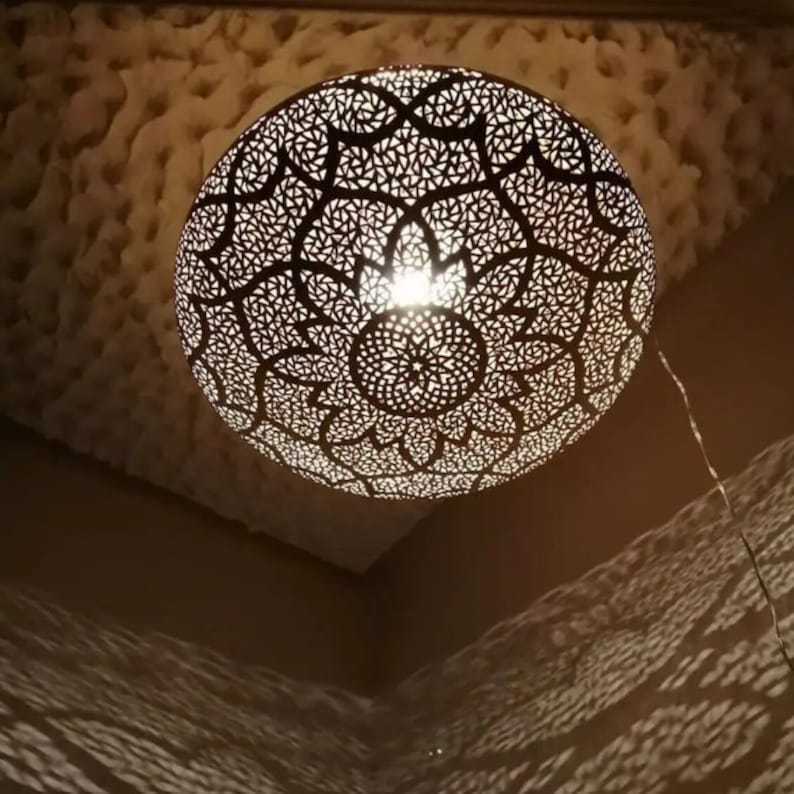 May include: A spherical, intricately patterned pendant light fixture. The metal shade features a detailed, openwork design, casting decorative shadows. The light source is visible within the fixture, and a cord hangs down. The fixture is suspended from a textured ceiling.