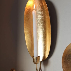 May include: Gold metal wall sconce with a candle holder. The sconce is shaped like a leaf and has a textured surface. A white candle is in the holder and is lit.