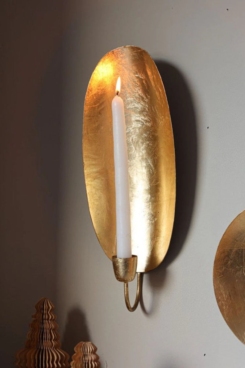 May include: Gold metal wall sconce with a candle holder. The sconce is shaped like a leaf and has a textured surface. A white candle is in the holder and is lit.