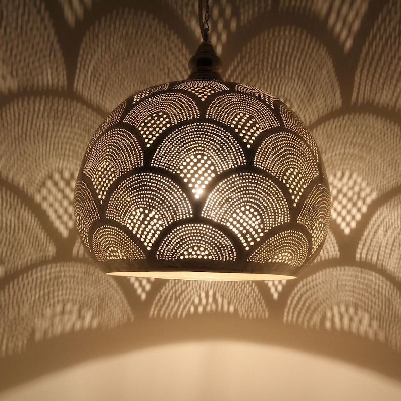 May include: A dome-shaped metal pendant light fixture with a dark finish, featuring an intricate pattern of small, punched-out circular holes arranged in overlapping semi-circular arcs that cast a matching shadow pattern on the surrounding area.