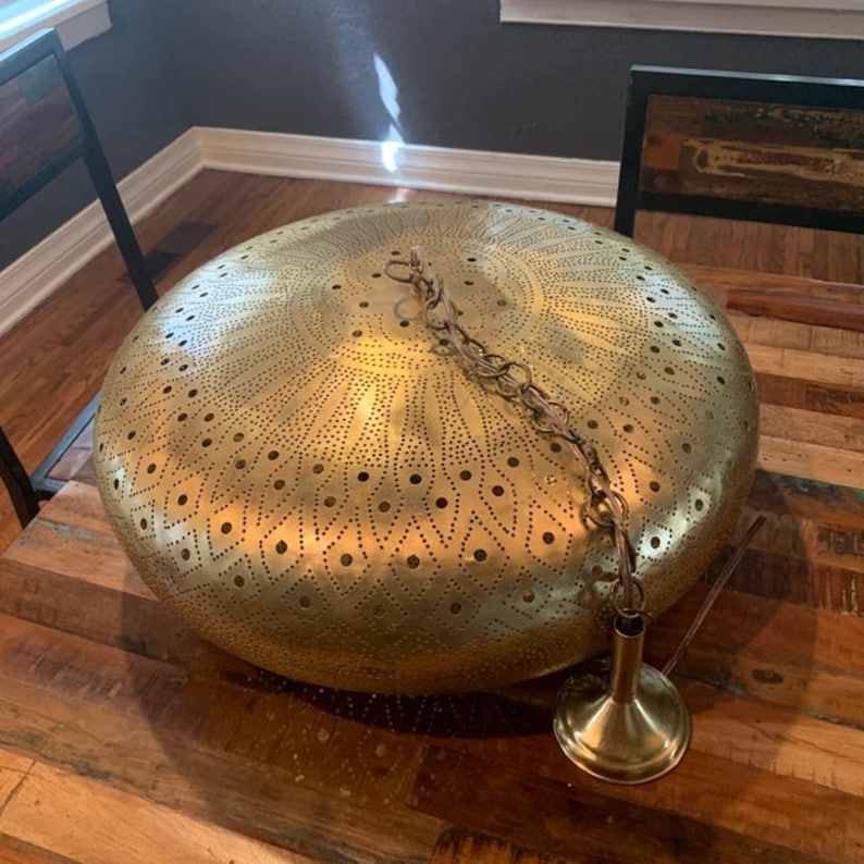Puede incluir: Una l&aacute;mpara colgante de metal dorado con una pantalla redonda perforada. La pantalla tiene un patr&oacute;n decorativo de c&iacute;rculos y l&iacute;neas. La l&aacute;mpara est&aacute; colgada de una cadena.