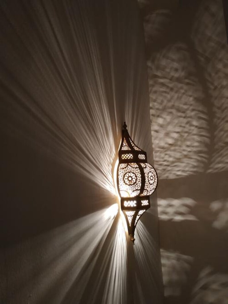 May include: A decorative metal lantern with intricate cutouts and a warm light shining through. The lantern is mounted on a wall and casts a soft glow on the surrounding area.